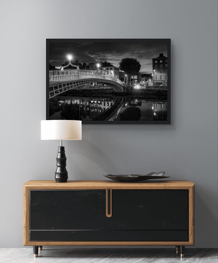 Black and white framed wall art print of Ha'penny Bridge in Dublin, Ireland at night. The iconic bridge is beautifully illuminated, with reflections shimmering in the River Liffey. Elegant, high-quality print