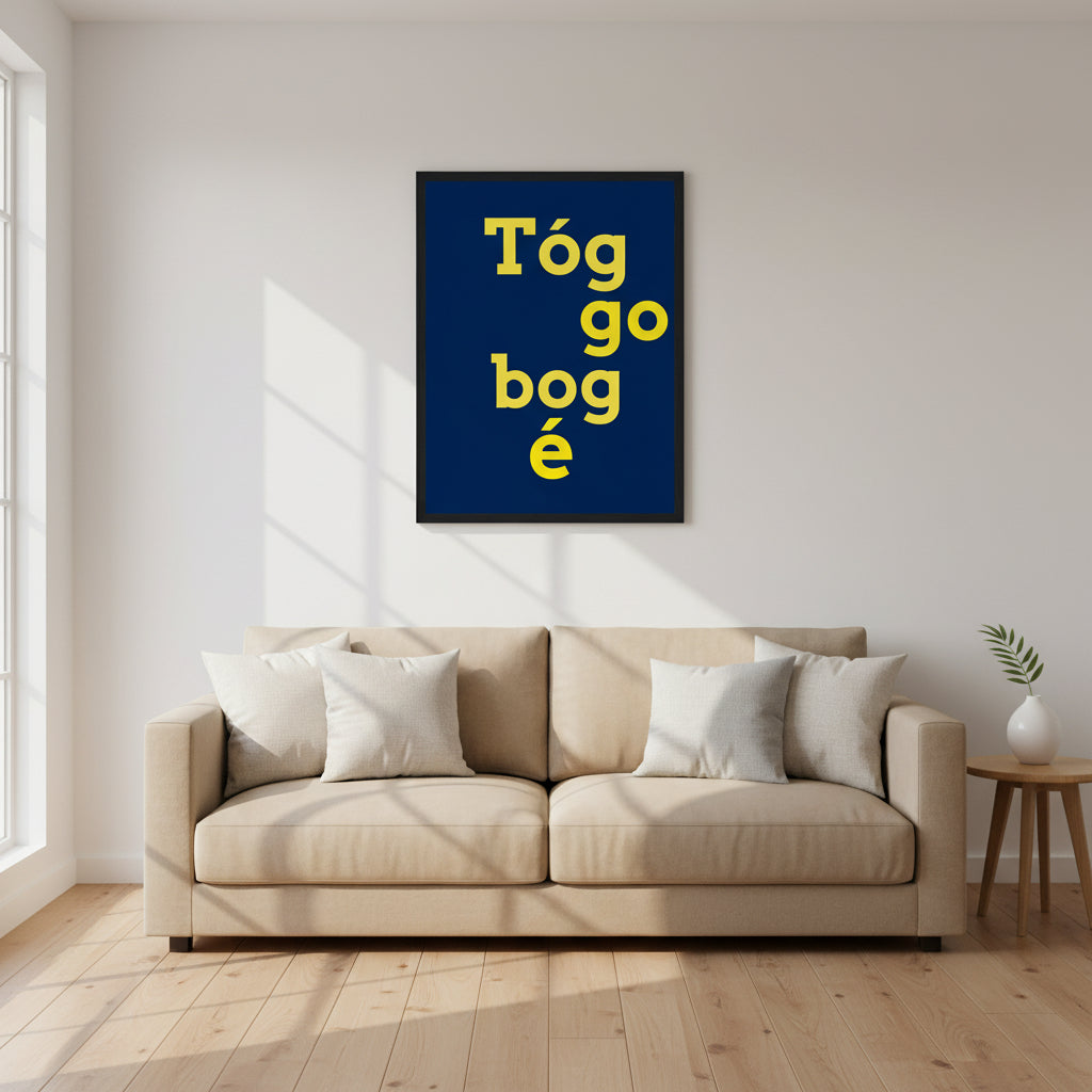 Framed “Tóg é go bog é” print in navy and yellow, centred above a beige sofa in a minimalist living room with white walls, oak floors, linen cushions and natural light.